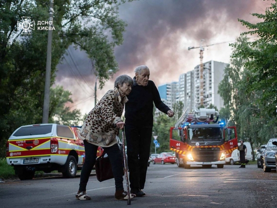 This handout photograph taken and released by the State Emergency Service of Ukraine on July 4, 2025, shows an elderly couple crossing a street as smoke rises in the background at a site of a strike following a mass Russian drone and missile attack on the Ukraine's capital Kyiv, amid the Russian invasion of Ukraine. Russia launched its largest-ever drone and missile attack on Ukraine overnight on July 4 just hours after a telephone call between US and Russian presidents ended without any breakthrough. (Photo by Handout / STATE EMERGENSY SERVICE OF UKRAINE / AFP) / RESTRICTED TO EDITORIAL USE - MANDATORY CREDIT "AFP PHOTO / State Emergency Service of Ukraine / HANDOUT" - NO MARKETING NO ADVERTISING CAMPAIGNS - DISTRIBUTED AS A SERVICE TO CLIENTS