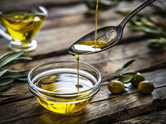 Pouring extra virgin olive oil from a vintage spoon to a glass container. Some olive branches comes from the left and right. The composition is on a rustic wooden kitchen table. Predominant colors are gold, green and brown. High resolution 42Mp studio digital capture taken with Sony A7rII and Sony FE 90mm f2.8 macro G OSS lens
