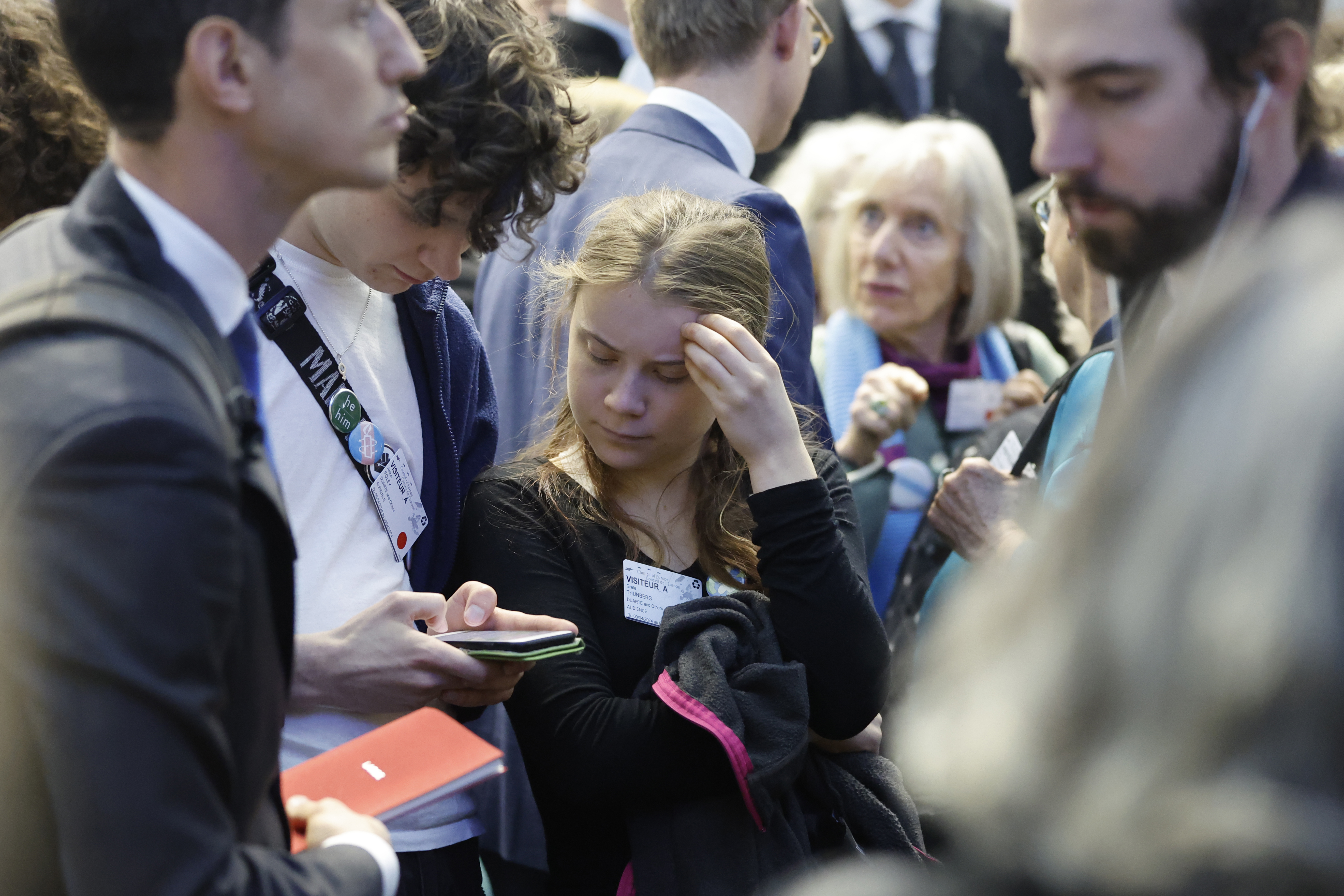 Swedish climate activist Greta Thunberg, center, watches a youth's phone inside the European Court of Human Rights, Tuesday, April 9, 2024 in Strasbourg, eastern France. Europe's highest human rights court threw out a case brought by six Portuguese youths aimed at forcing countries to meet international obligations to reduce greenhouse gas emissions, but sided with a group of senior Swiss women who also sought such measures. (AP Photo/Jean-Francois Badias)